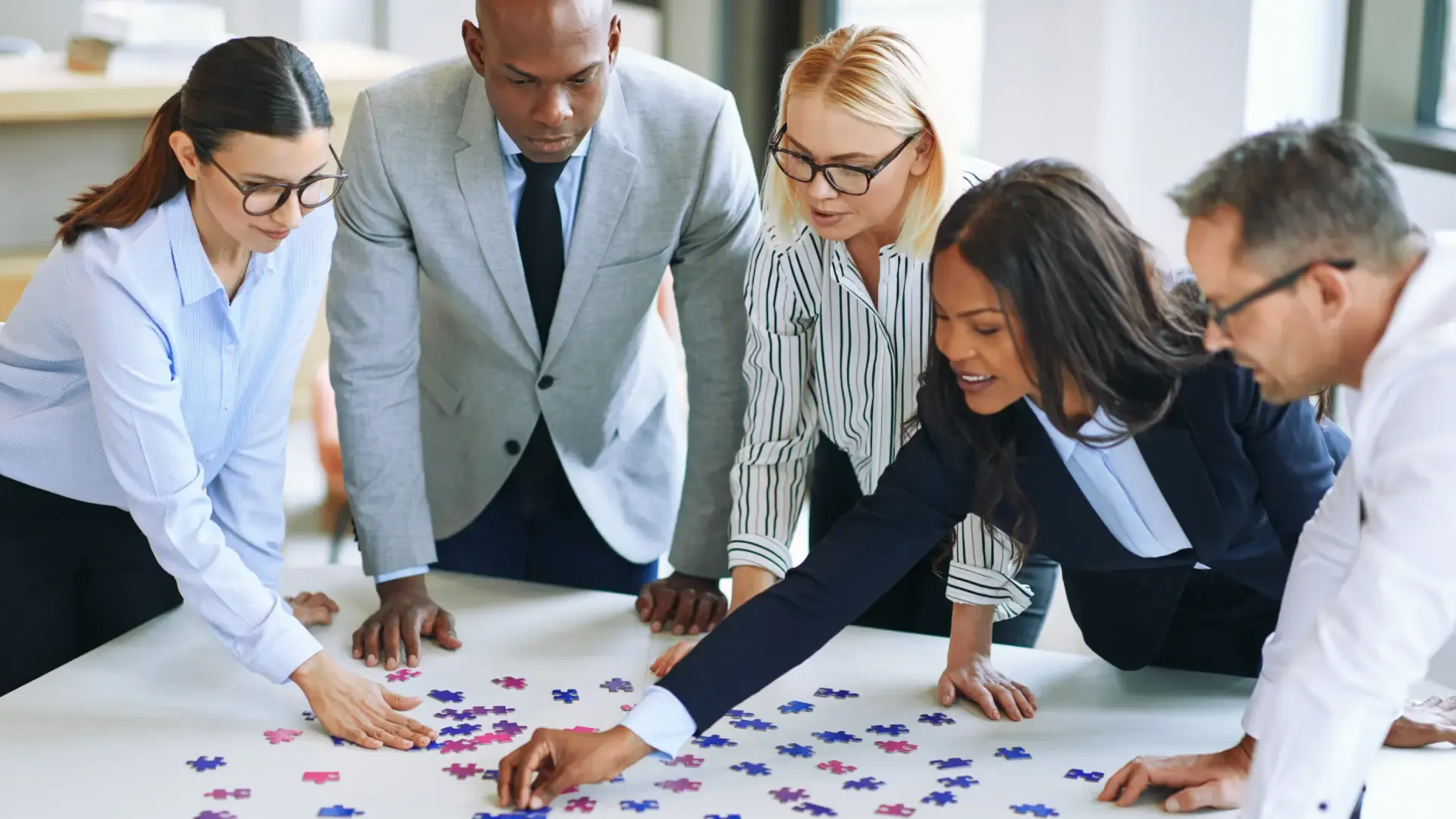 Team legt auf einem Tisch Puzzleteile zusammen: Wissen liegt verteilt und muss erst zusammengeführt werden